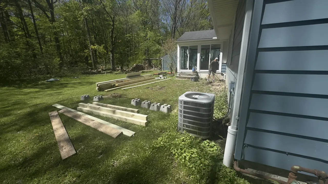 Pressure-treated lumber staged on the lawn beside a blue house before deck framing.
